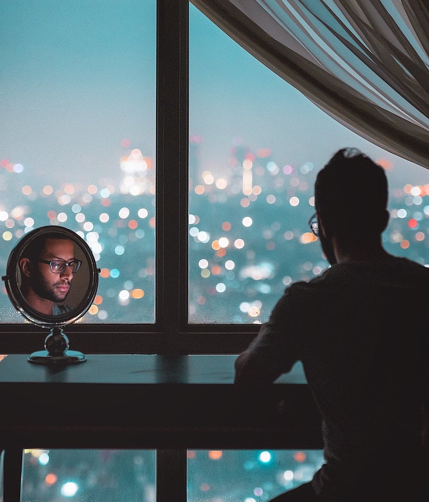 comfort zone - personne de dos assise devant une fenêtre avec une vue sur une ville dont on voit le visage dans un petit miroir