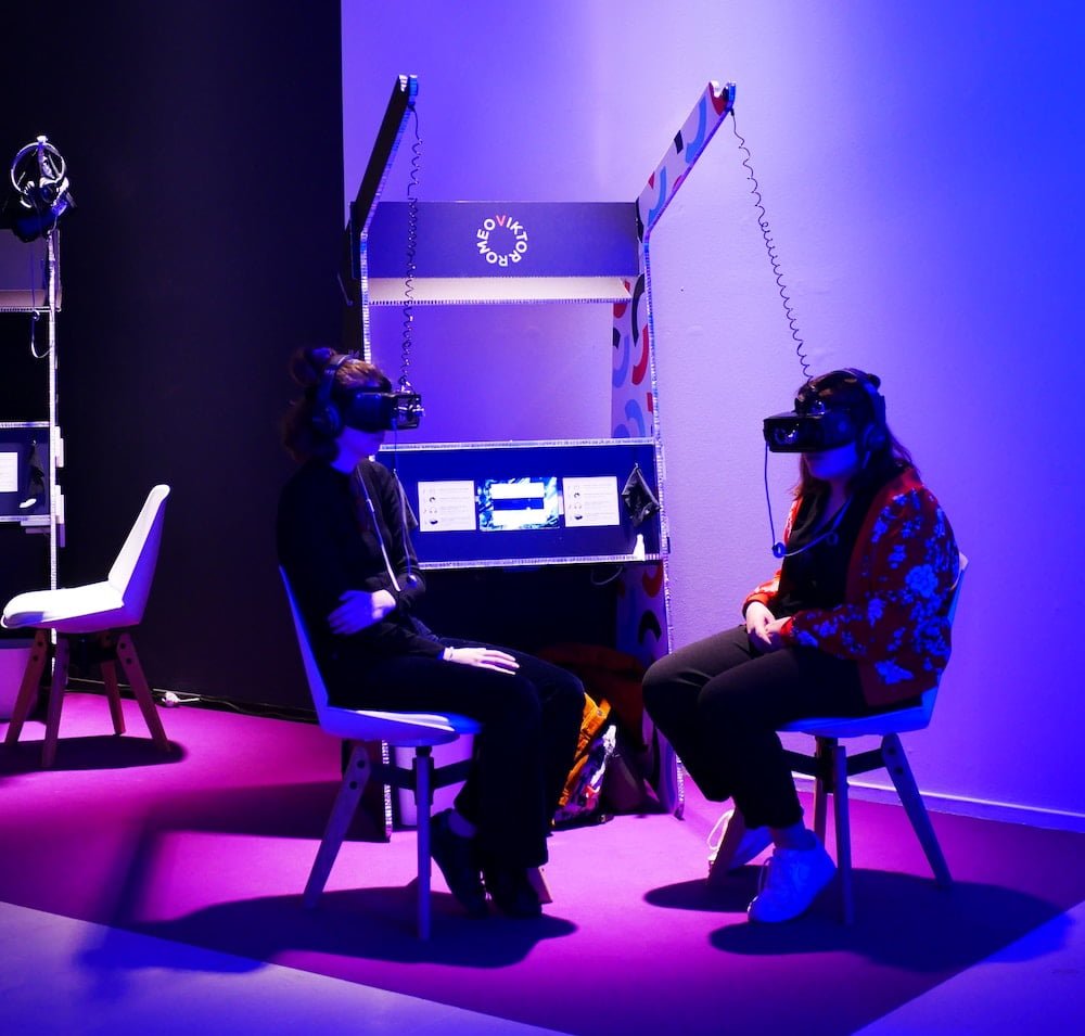 Deux personnes assises sur des chaises avec un masque de réalité virtuelle dans une lumière mauve (nouvelles technologies)