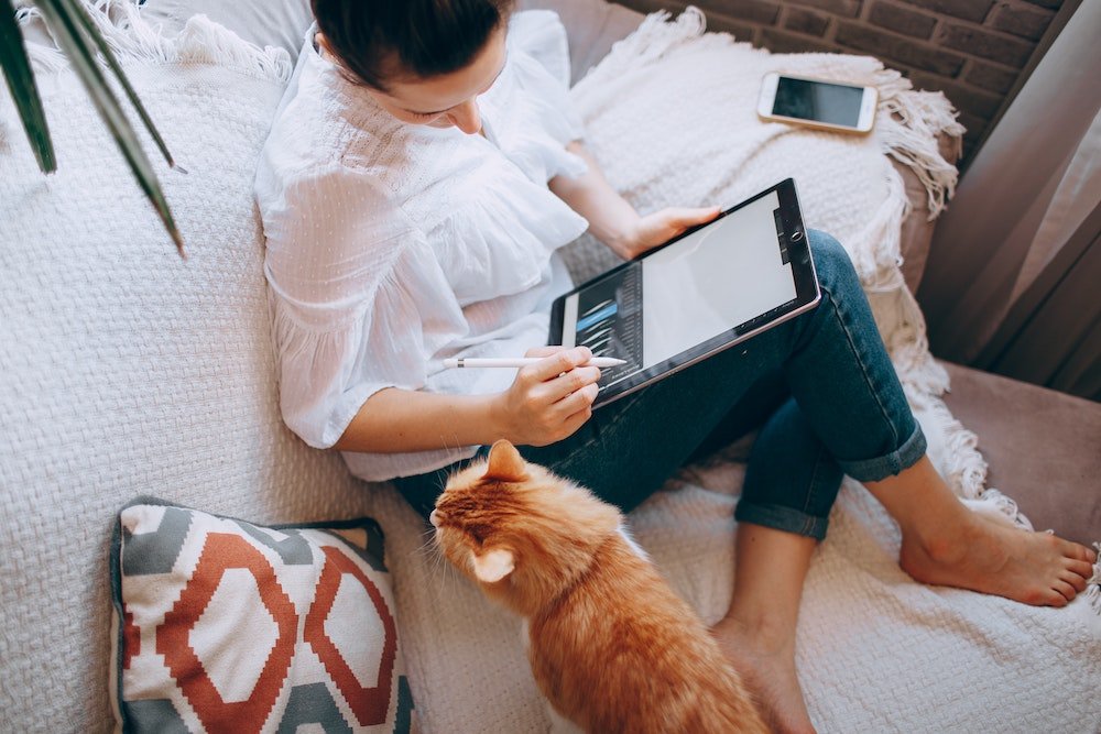 Pénurie | Femme qui dessine sur une tablette assise sur un divan avec un chat